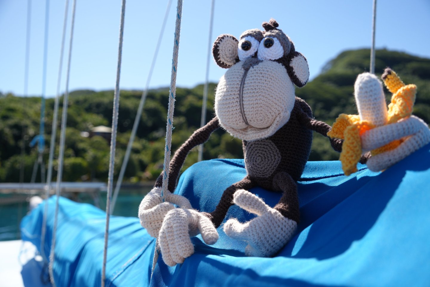 A handmade crochet monkey plush toy holding a banana on a blue boat sail with a tropical island background.