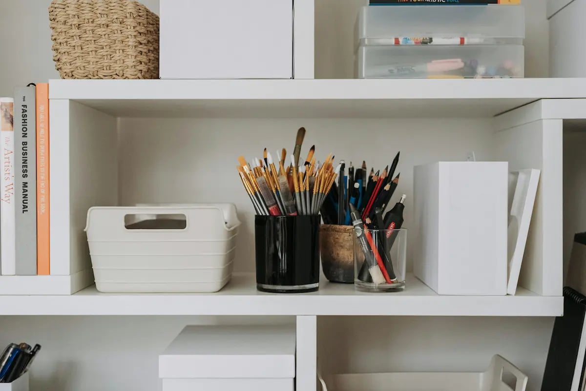 Organized art studio white bookshelf featuring paintbrushes, pencils, and craft storage bins.