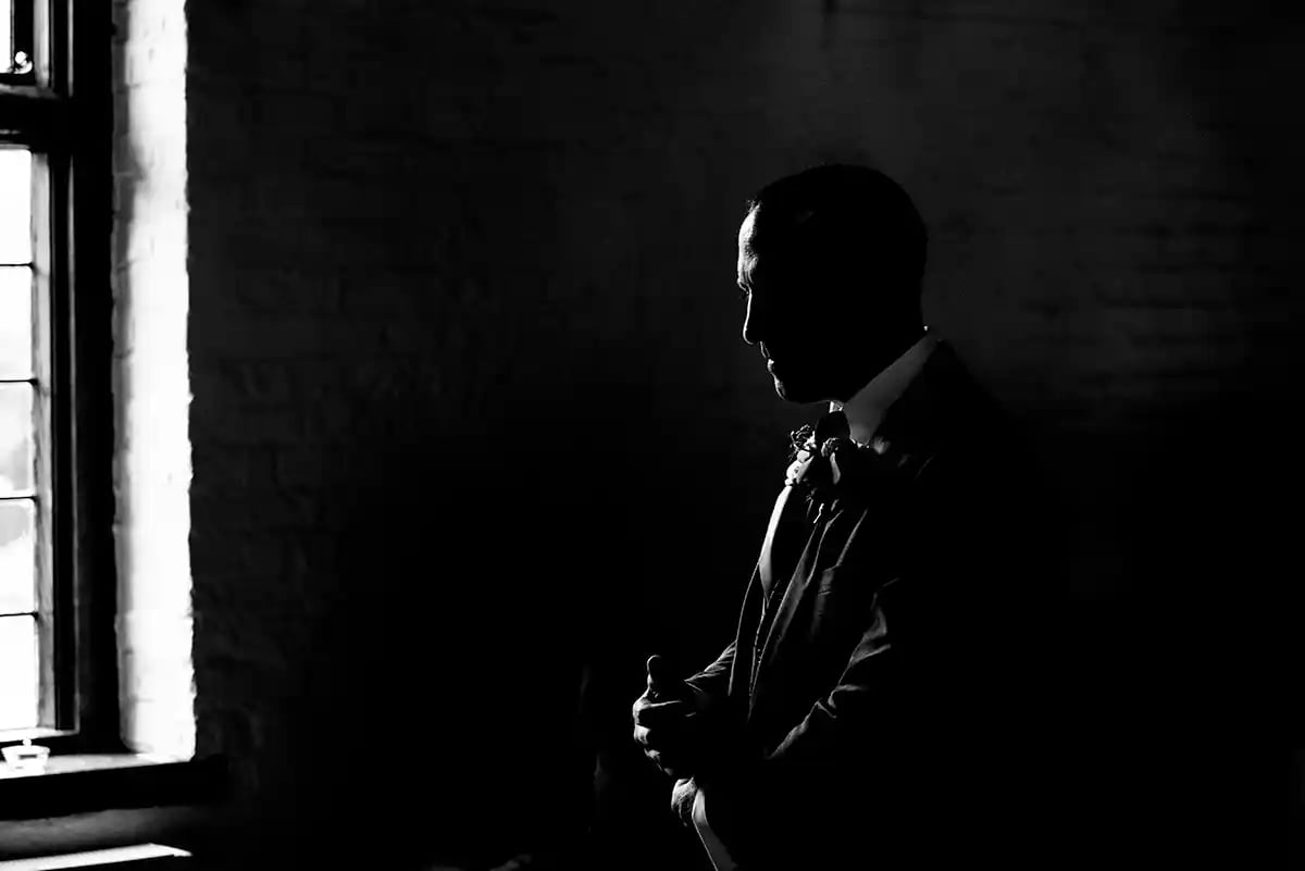 Black and white silhouette of a groom in a tuxedo standing by a window before a wedding ceremony.