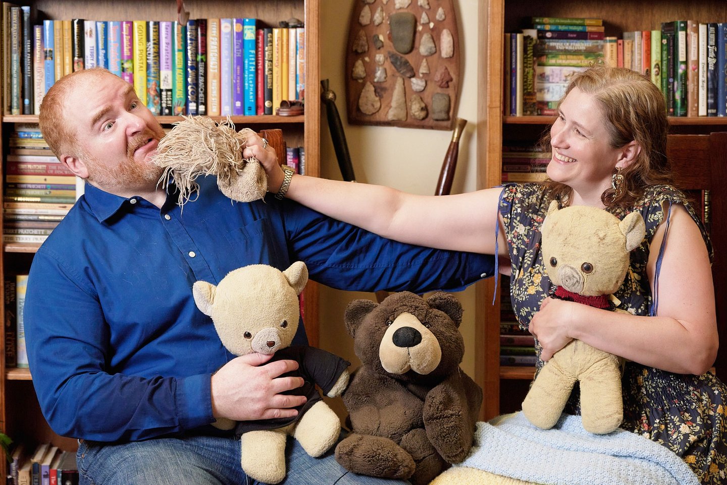 a man and woman sitting on a chair with teddy bears fighting each other