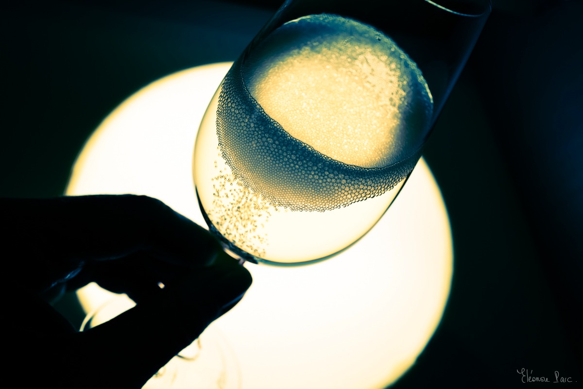 Abstraction Vinicole – Flûte de vin effervescente, couronne de bulles, main en ombre