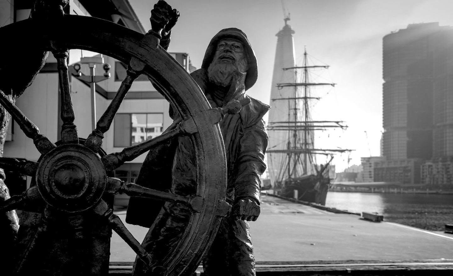Black and white statue of a sailor at a ship's wheel overlooking a harbor with a tall ship.