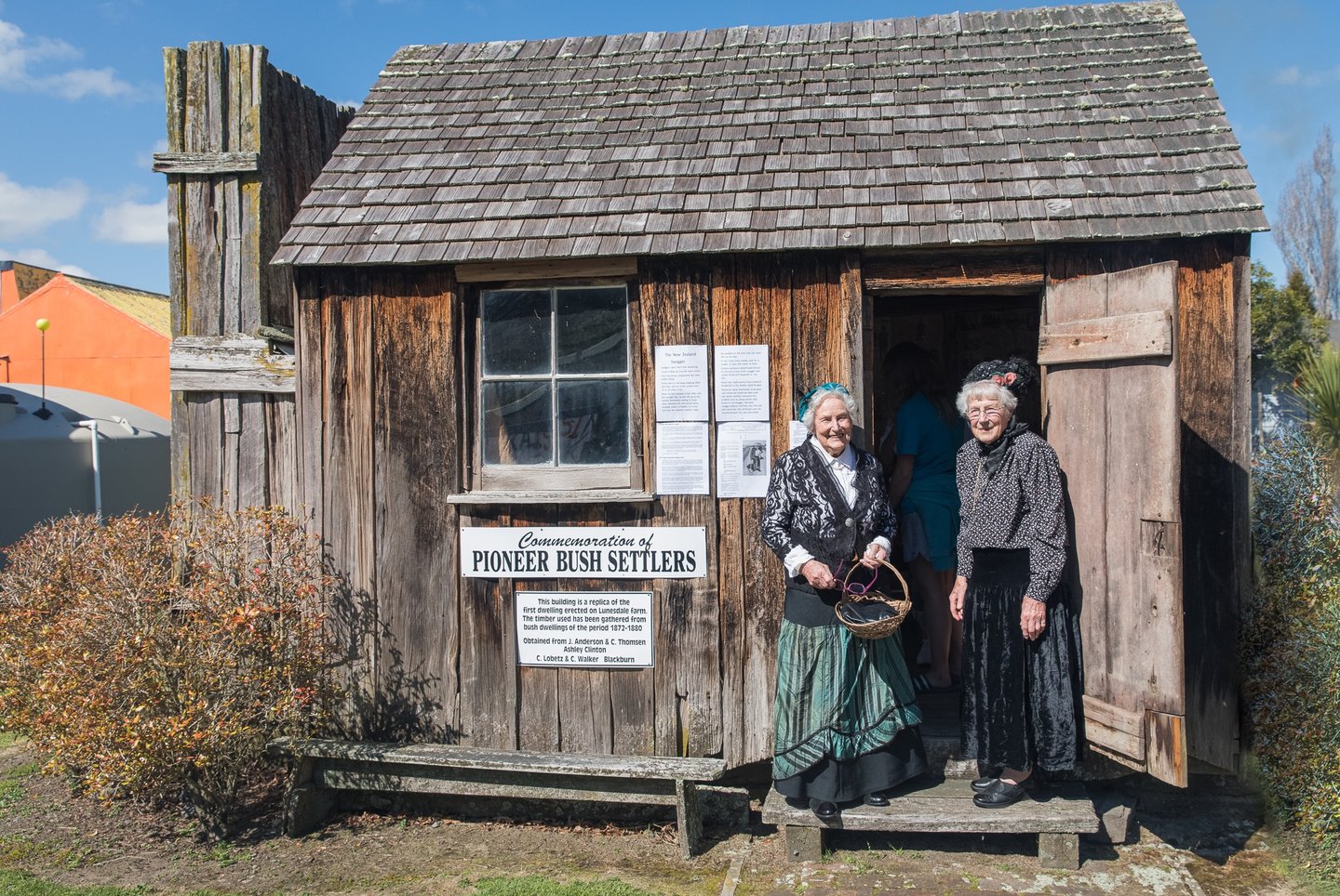 two women in period costumes standing in front of a traditional Pioneer Bush Hut