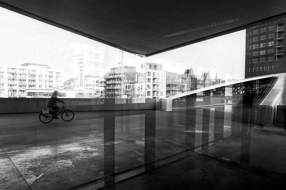 Black and white street photography of a cyclist riding past modern urban architecture and reflections.