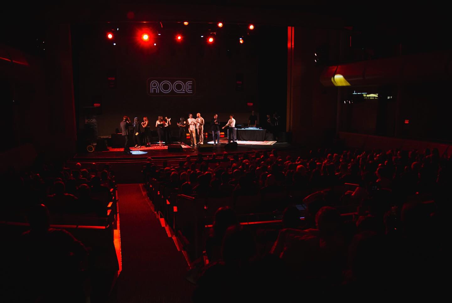 A large audience watches a live event on stage with red theater lighting
