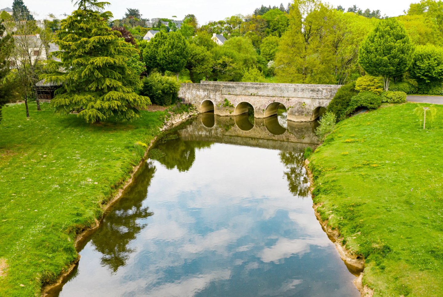 Pont historique en pierre se reflétant dans une rivière calme, entouré d'arbres et d'herbes verdoyan