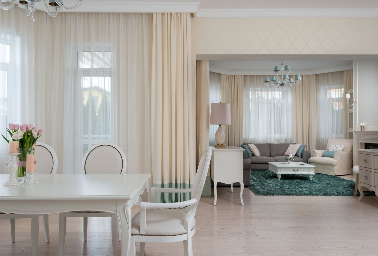 Elegant open concept living and dining room featuring white furniture, soft cream curtains, and a teal area rug.