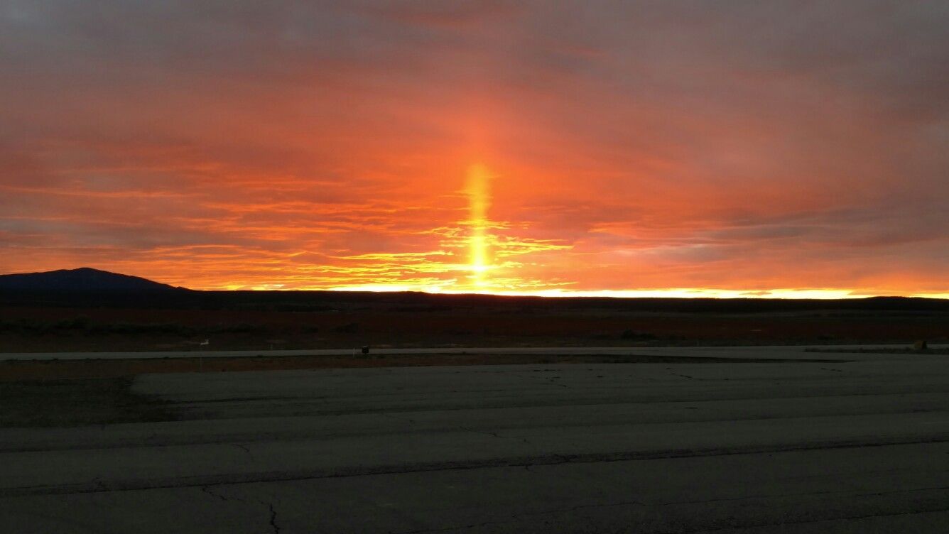 A vibrant orange sunset over a dark landscape featuring a rare vertical sun pillar light phenomenon.