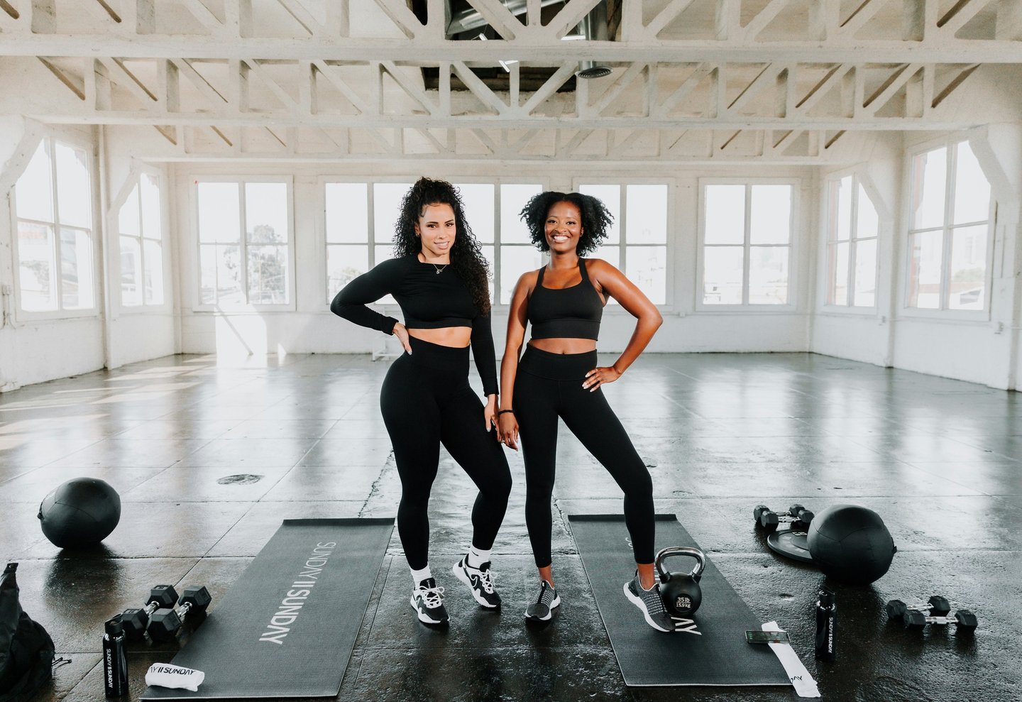 Two smiling women in black activewear standing on yoga mats in a bright fitness studio with gym equipment.