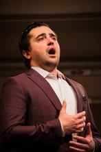 A male opera singer in a suit, mouth wide open, projects his voice with his hands up to his chest