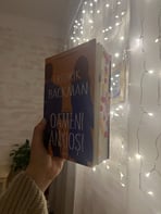 Mână ținând cartea 'Oameni anxioși' de Fredrik Backman pe un fundal de luminițe de basm.