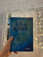 Mână ținând cartea 'Flori pentru Algernon' de Daniel Keyes pe un fundal de luminițe de basm.