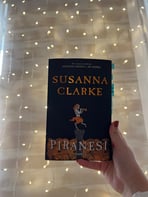 Mână ținând cartea 'Piranesi' de Susanna Clarke pe un fundal de luminițe de basm.