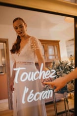 Une femme habillée en robe de mariée touche l'écran tactile du miroir photobooth de pixtoevent