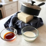 Tofu wrapped in a towel under a heavy pan; bowl with whisked soy sauce mixture