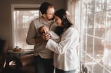 Mom holding and looking at their newborn while dad kisses her head in front of a window during a Gra