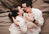 Mom and dad cuddling in bed with their newborn, looking at each other lovingly from above.