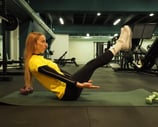 Una donna in maglia gialla esegue l'esercizio 'boat pose' su un tappetino da yoga in palestra, concentrandosi sull'equilibrio