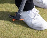 Person picking up a golf tee of the grass using the TeeZUP tool on grass, with a club and white sneakers visible.