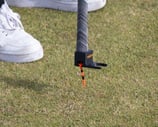 A golfer uses the 'TeeZUP' tool, attached to the end of a golf club, to pick up a orange tee in place on the grass