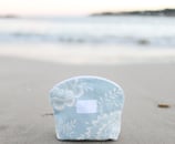 Trousse de maquillage ÉTOFÉ en tissu recyclé bleu clair à motifs floraux, posée sur une plage