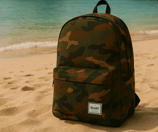 a Herschel camo backpack sitting on the sand of a beach in Thailand