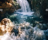 a waterfall in the middle of a river with sun shining through the trees