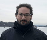 Photo of a man wearing glasses, with a slight smile. Behind him is the sea, a lighthouse and a forest. It is cloudy.