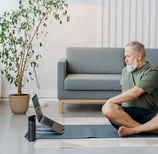 Hombre adulto mayor sentado con las piernas cruzadas sobre un tapete de yoga en su sala.
