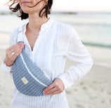 Femme debout à la plage portant un sac banane ÉTOFÉ bleu en tissu revalorisé, tenue blanche estivale