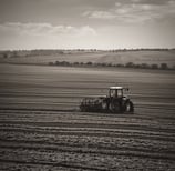 A red tractor with a large attached irrigation reel is positioned on a grassy path in a field landscape. The background consists of dense, dark green trees, while the foreground displays cultivated fields with patches of green and brown earth.