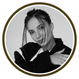 A person in a stylish black outfit poses with one arm wrapped around themselves against a muted backdrop.