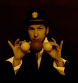 a man in a suit and hat with two hands up to the camera holding juggling balls