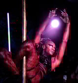 black woman suspended by mesh cover legs on pole, arms outstretched gracefully backlit by spotlight