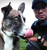 smiling man with glasses and black baseball cap and dog licking its face