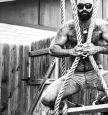 hairy, shirtless bearded man in sunglasses and cutoff shorts hugging ropes