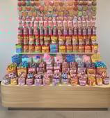 A candy and snacks kiosk in a shopping mall features a variety of colorful candies and packaged snacks. A customer, wearing a scarf and carrying a shopping bag, is selecting items from the display while the vendor, dressed in a green uniform, stands on the opposite side. Other shoppers are visible in the background, walking past the kiosk.