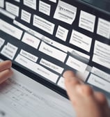 A person is signing documents on a dark wooden table. Multiple sheets of paper are spread out, with text and tables visible on them. The person is holding a black pen and wearing dark clothing, creating a contrast with the white papers.