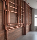 A large, brown metal pipe is installed along the corner of a room. The pipe is supported by several bands of white tape or insulation material. Adjacent to the pipe, a white cloth or towel is hanging on a hook. The walls and ceiling are white, with a textured ceiling pattern, and there are visible cables or wires near the pipe.