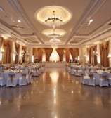 An outdoor wedding setup features neatly arranged white chairs facing a wooden arch adorned with white fabric and floral decorations. The setting overlooks a calm body of water, surrounded by lush greenery and a well-kept lawn.