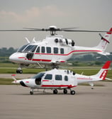 A white helicopter is parked on a grassy field with dense green trees and a wooden cabin in the background. The rotor blades are stationary, and the helicopter bears a logo and registration markings on the side.