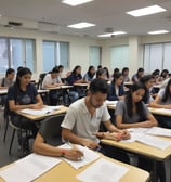 A group of children sitting at wooden desks in a classroom environment. They are focused on writing with pens, and wearing green uniforms. The room appears well-lit with natural light coming through a window.