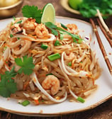 A plate of pad thai garnished with green onion, crushed peanuts, and lime wedges is on a blue plate. It includes rice noodles, pieces of chicken or tofu, and is sprinkled with red chili flakes.