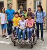 Several adults are gathered around a young girl, placing their hands gently on her shoulders in a supportive gesture. They seem to be outdoors, with trees and a small building visible in the background. The scene conveys a sense of care and community.