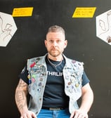 man in goatee with denim vest covered in pins with cartoon angel and demon on black background