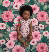A baby lies on a floral-patterned fabric, wearing patterned shorts. Next to the baby is a doll with a floral dress and striped leggings. The background features pink flowers and large leaves, creating a playful and cozy setting.