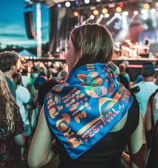 A vibrant crowd at an outdoor music festival at night, focusing on a young woman wearing a polyester scarf with printed the l