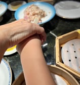 一隻手握住另一隻手嘅手腕，背景係餐桌，上面有碟子同竹蒸籠，場景似係飲茶或點心餐廳。