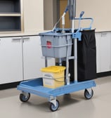 A cleaning cart is positioned in an outdoor area with a concrete building structure in the background. The cart holds a large bucket and cleaning supplies, including a striped cloth and a pink cloth hanging over the handle. A green scrubbing brush is placed on the bottom platform. Surrounding the area are vibrant flower beds with yellow and white flowers, as well as bushes and trees, contributing to a tidy and well-maintained environment.