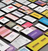 A collection of various office supplies and packages are scattered across a table. Items include a red phone, a serrated knife, rolls of packing tape, scissors, markers, a stapler, and several cardboard boxes. The background features a stack of green material, possibly paper or fabric, and a window with blinds.